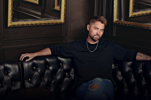 Brett Young wearing a black shirt, gold chain and ripped blue jeans. Sitting on a tufted black leather corner booth in a luxe wood paneled room.